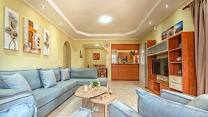 A living room with a sofa, coffee table, and TV stand. There are paintings on the wall and a kitchen in the background. - Villa Eleon with private pool, BBQ and beautiful garden. (Zakynthos)