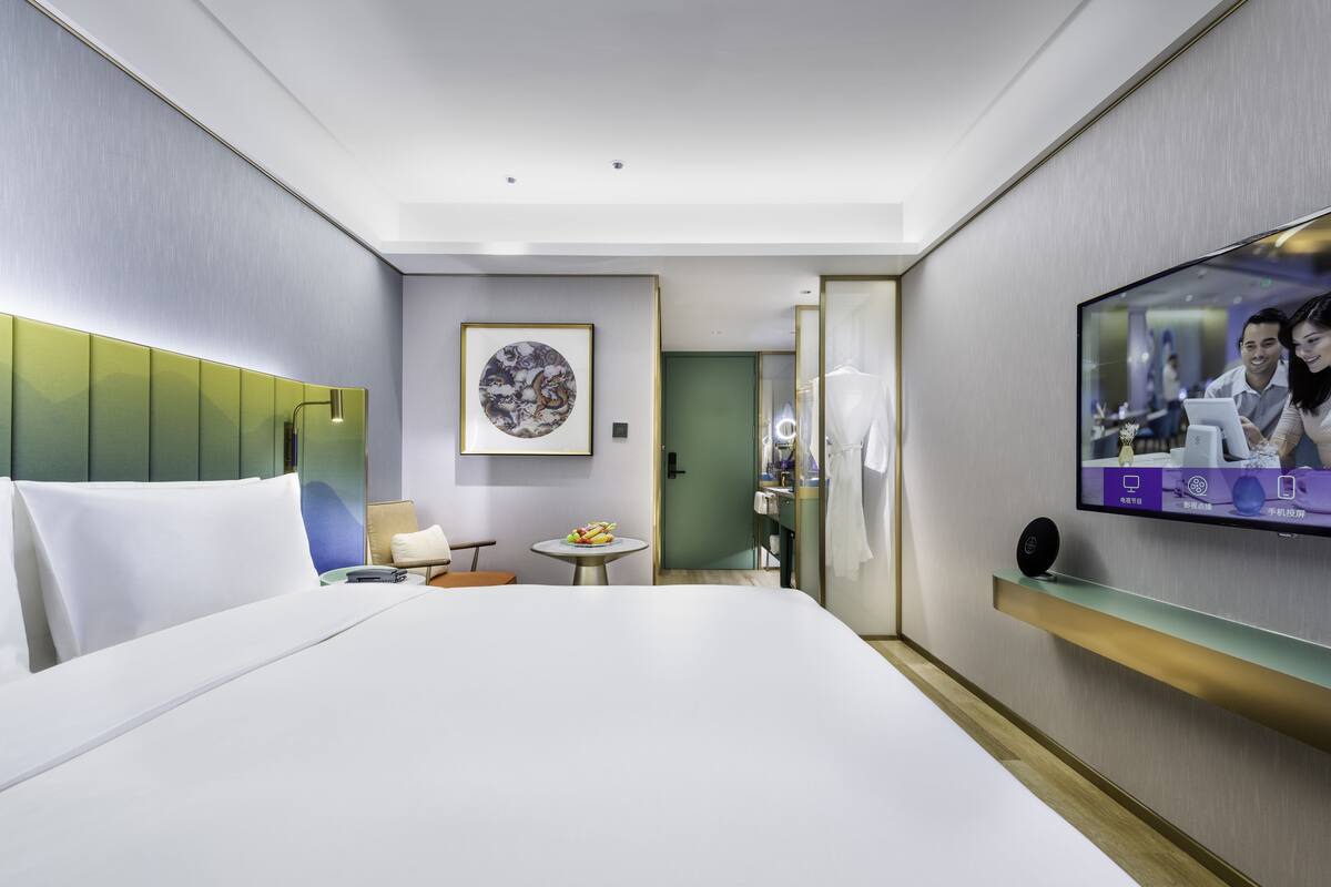 Een moderne hotelkamer met een groot bed, een klein tafeltje met een fruitschaal, een televisie aan de muur en een spiegel.