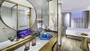 Una habitación de hotel moderna con un espejo grande, un tocador con artículos de aseo, una cama con cabecero blanco y una mesita con una bandeja de frutas.