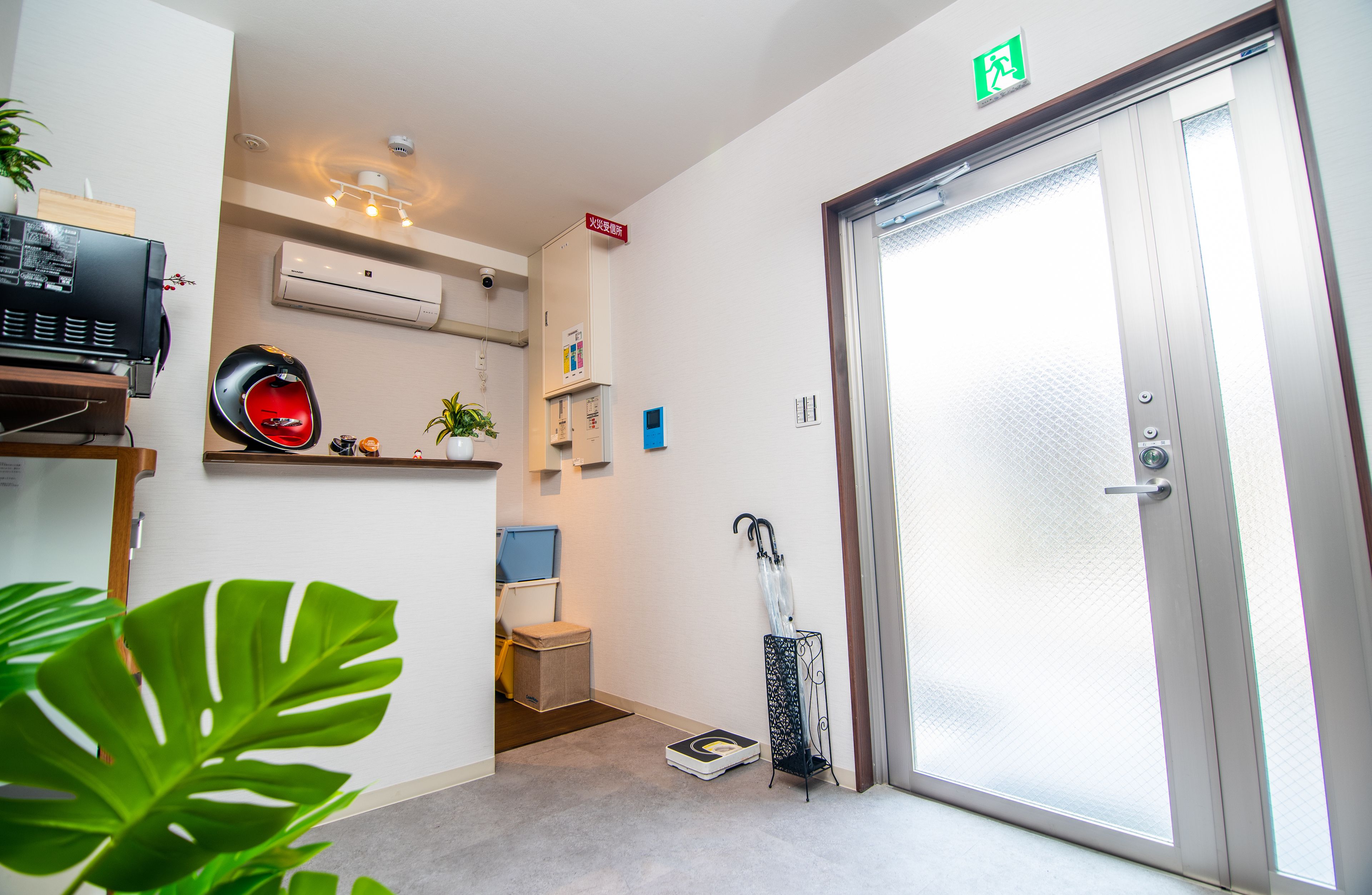 A hotel room with a door, a scale, a plant, a shelf with a helmet, and an air conditioning unit.