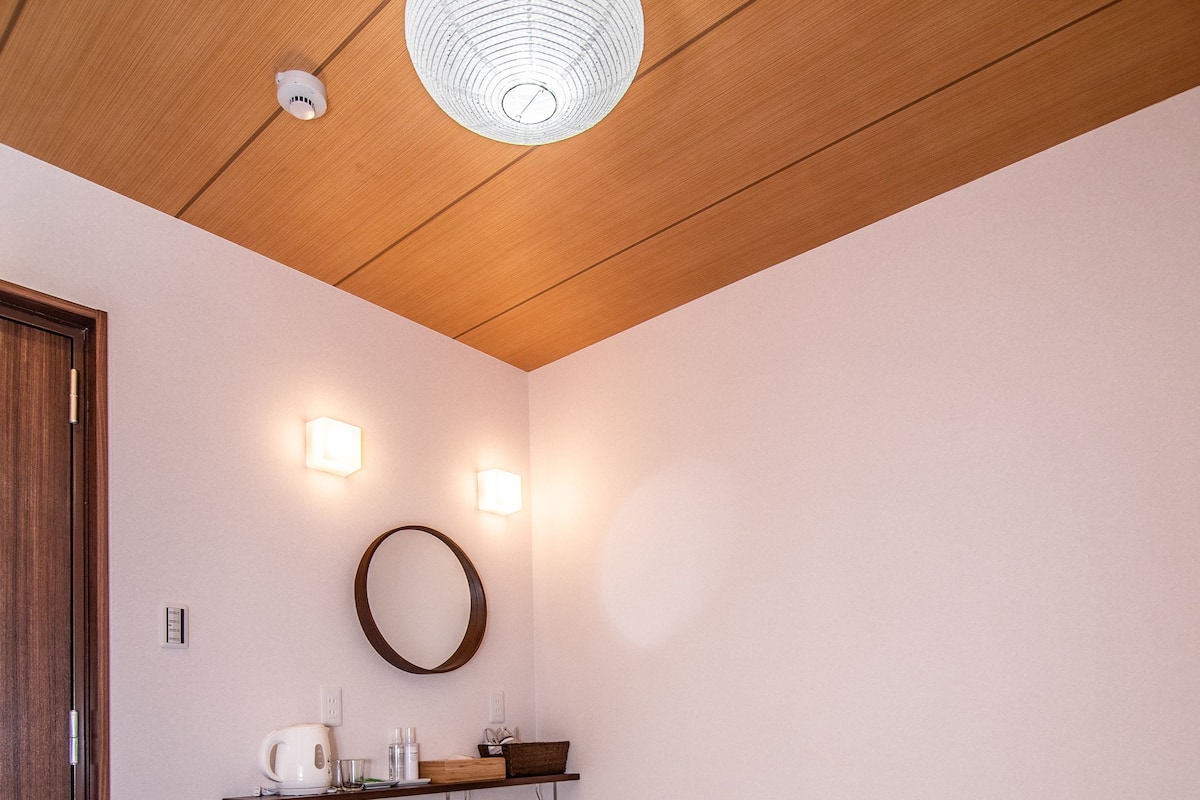 A traditional Japanese room with tatami flooring, a low wooden bed, a small wooden cabinet, and a wall-mounted shelf with a kettle and glasses.