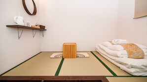 A room with tatami flooring, a bed with white bedding, a wooden stool, a shelf with toiletries, and a wall-mounted mirror. - Kujira 202 (Osaka)