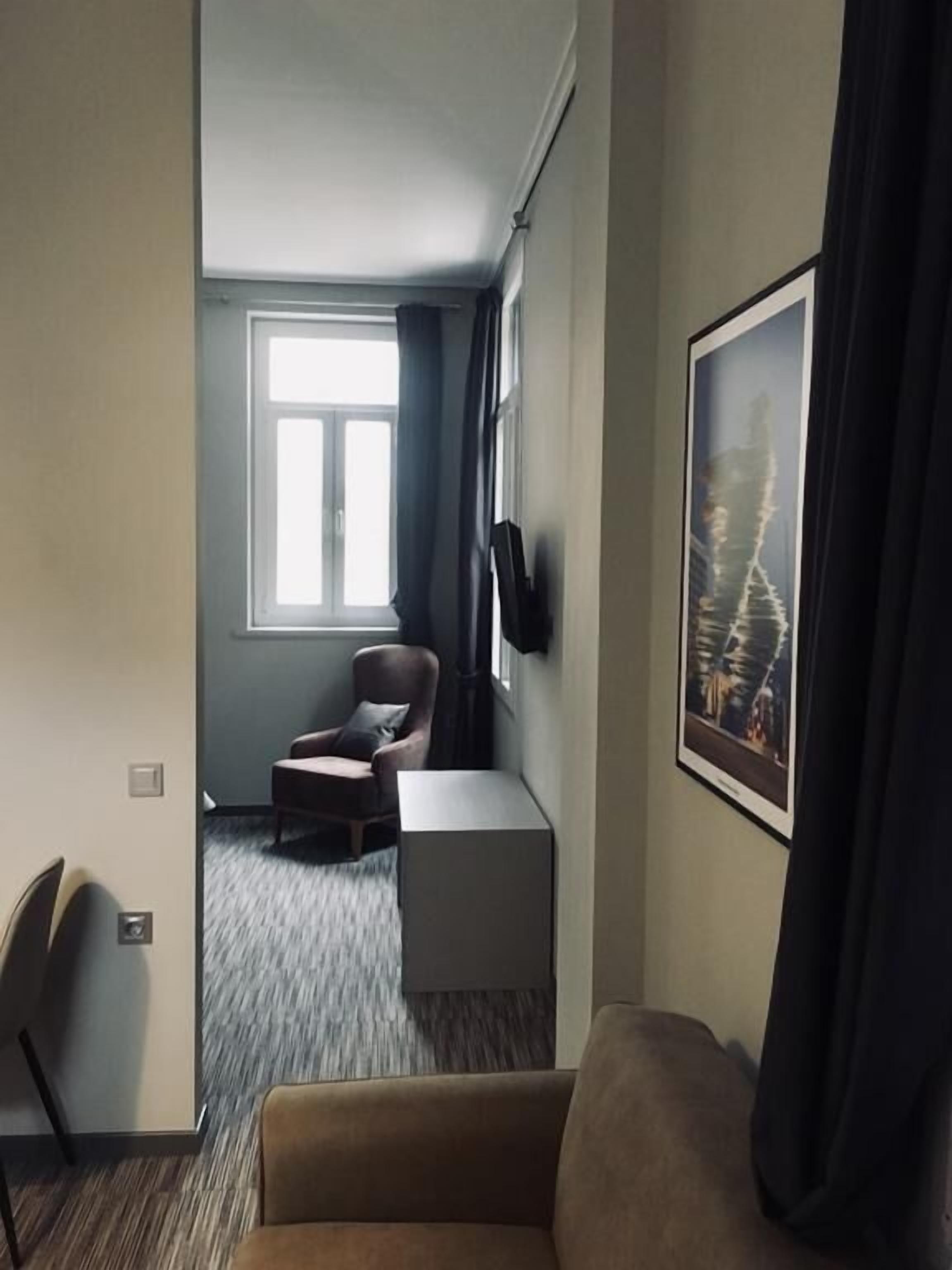 Une chambre d’hôtel avec un canapé, un fauteuil, une petite table, un téléviseur fixé au mur et une fenêtre avec des rideaux.