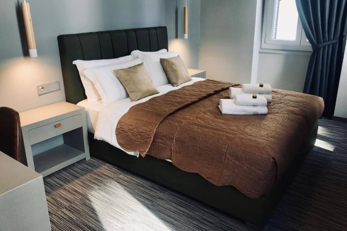 A neatly made bed with a brown blanket and white pillows, a bedside table with a lamp, and a window with curtains.