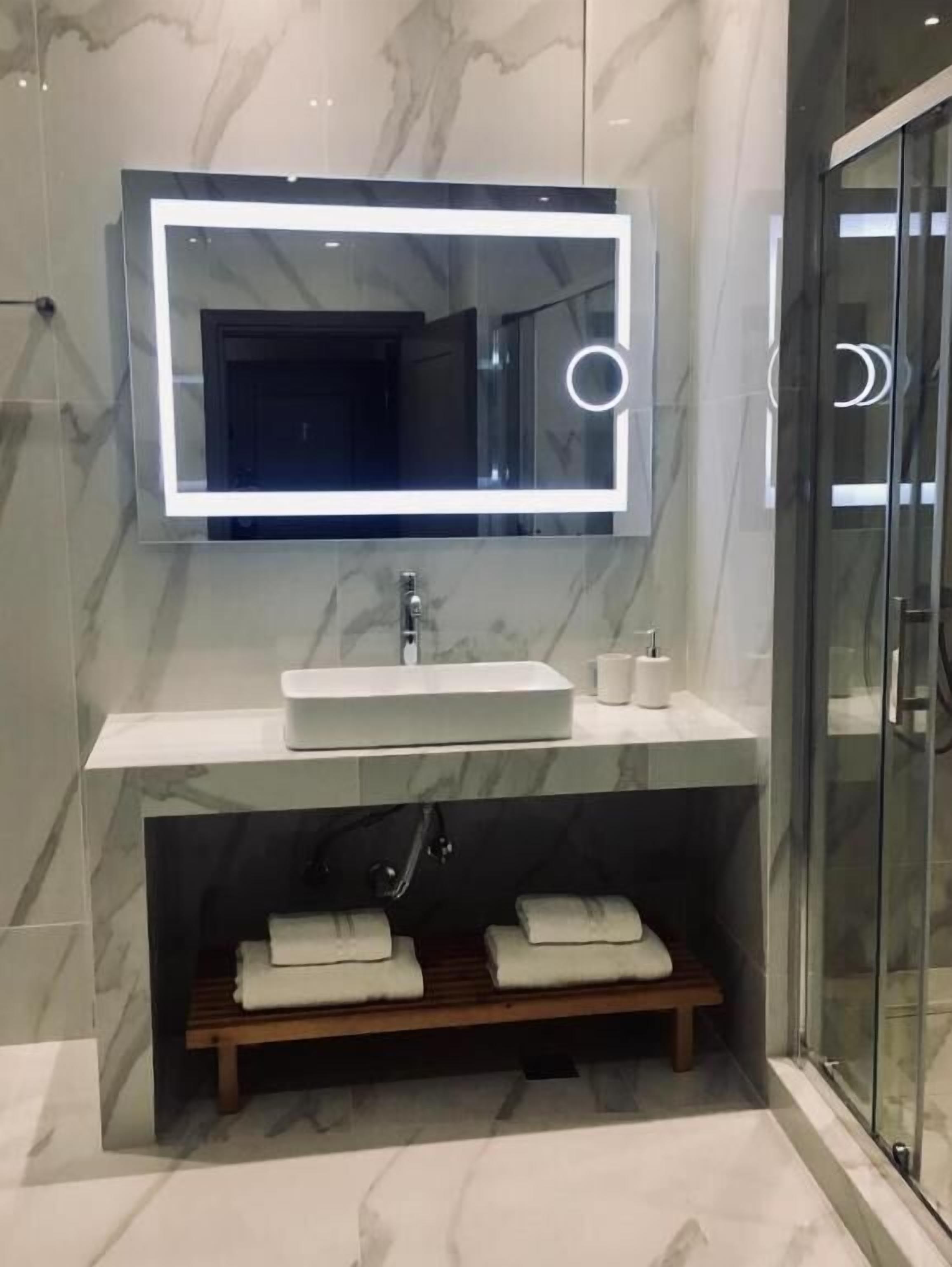A modern bathroom with a marble wall, a large mirror with integrated lighting, a white sink, and a glass shower enclosure.