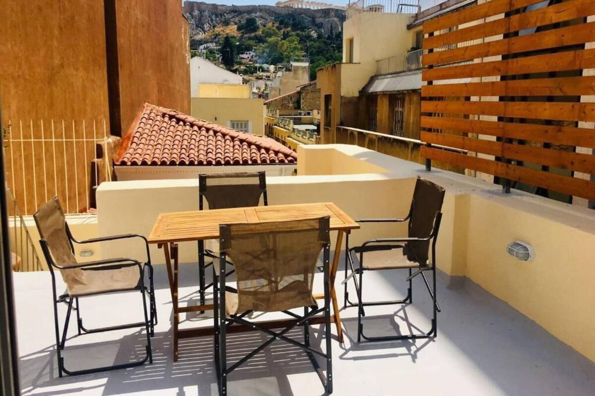 A rooftop terrace with a wooden table and chairs, a view of a cityscape, and a wooden fence.
