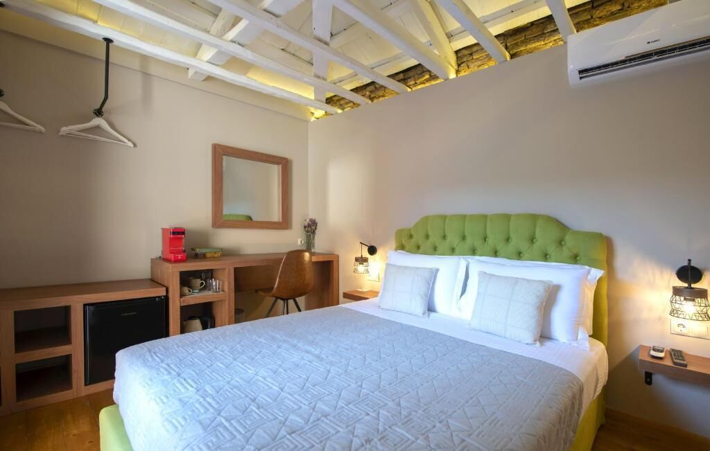 A bedroom with a green upholstered headboard, a bed with white linens, a desk with a red coffee maker, a chair, and a small bedside table with a lamp.