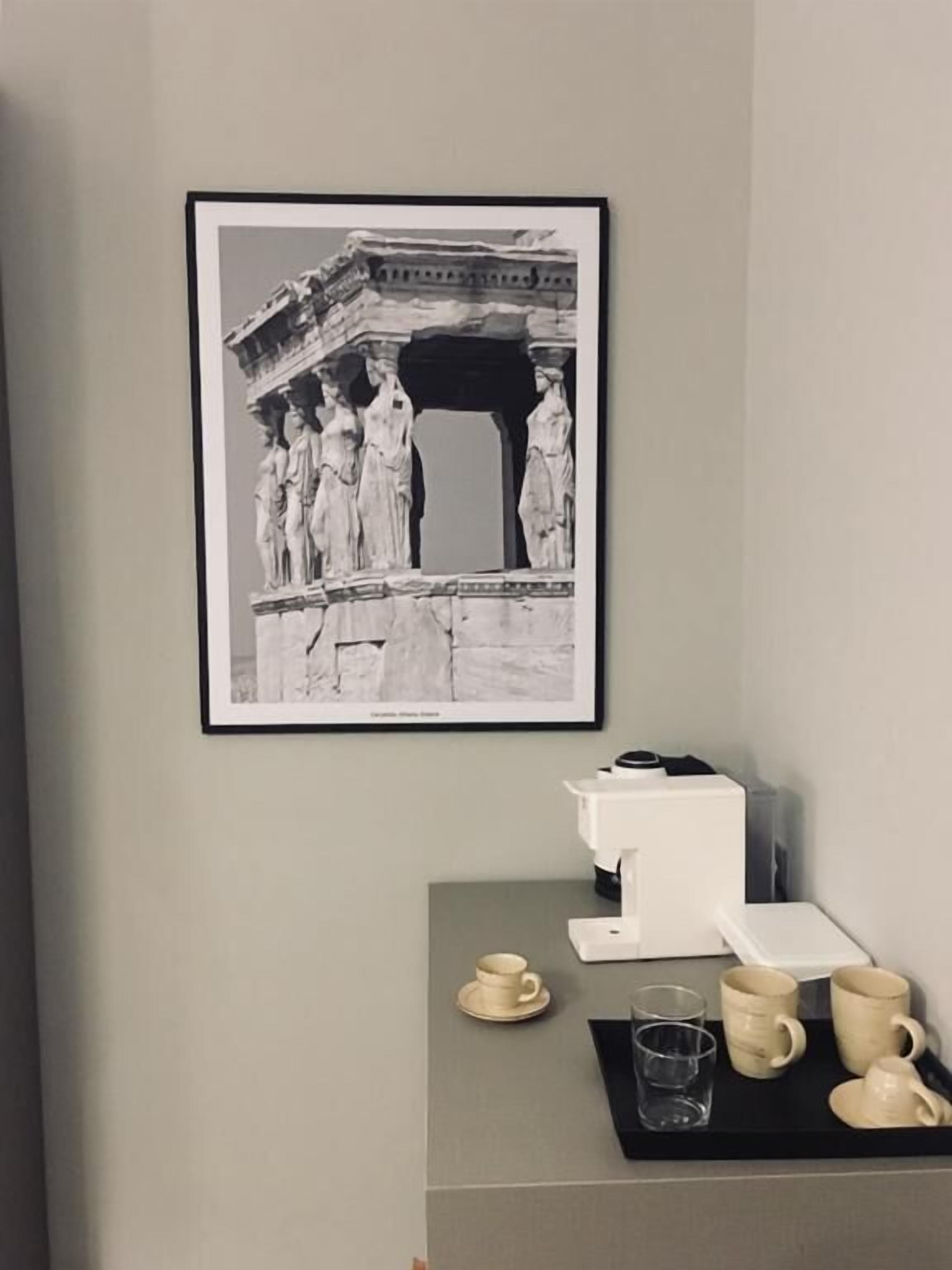 Fotografia in bianco e nero incorniciata che ritrae statue greche antiche su una parete, una macchina da caffè e un vassoio con tazze e un bicchiere.