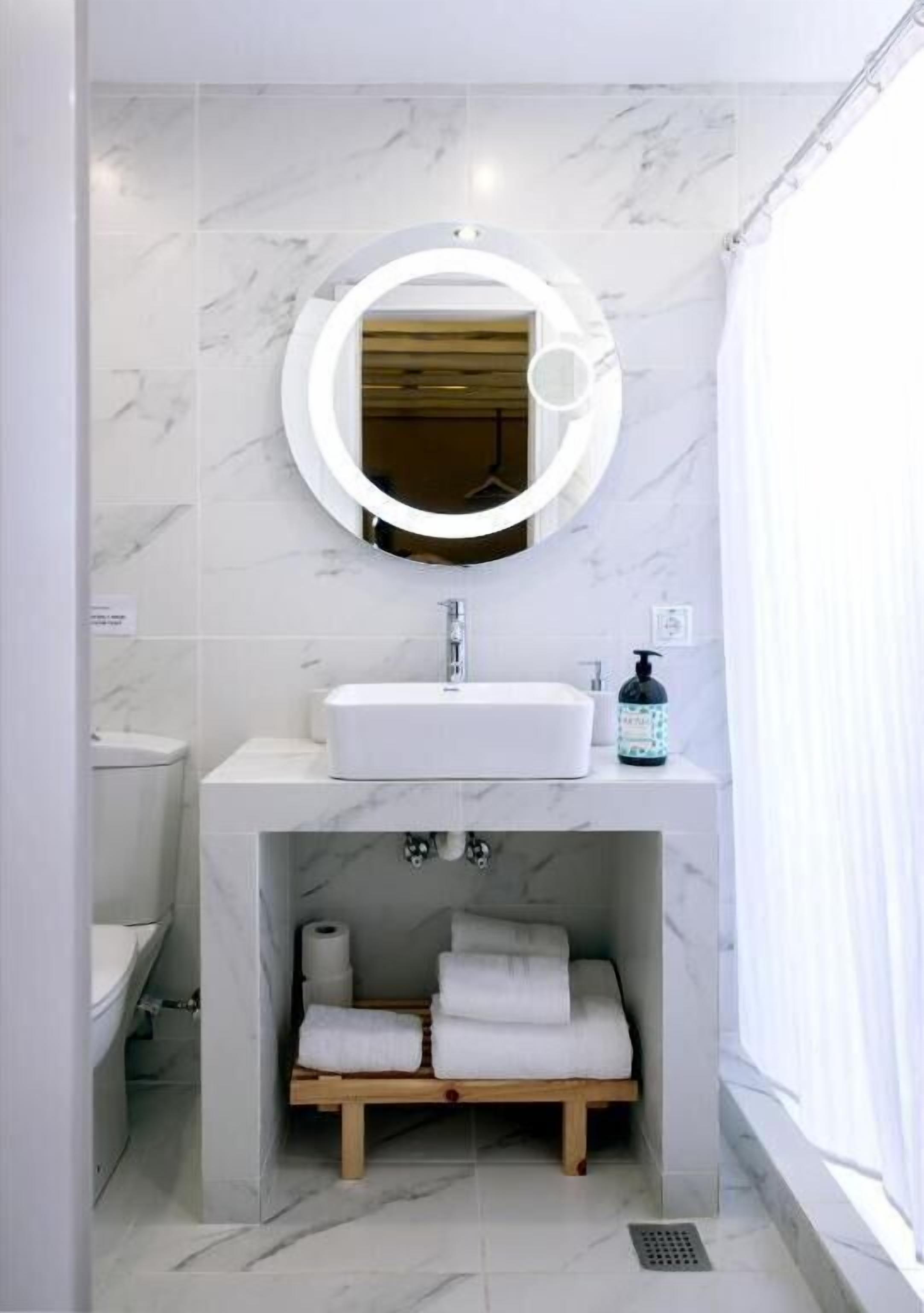 Une salle de bain avec un lavabo en marbre blanc, un miroir rond avec un éclairage intégré et une étagère en bois où sont posées des serviettes.