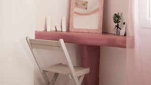 A bathroom with a pink vanity, a mirror, a small shelf with toiletries, and a foldable chair. - Pasithei Studios (Santorini)