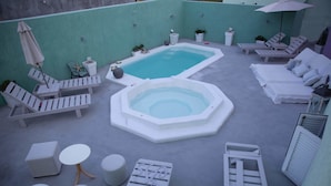 A rooftop pool area with lounge chairs, a hot tub, and small tables. - Pasithei Studios (Santorini)