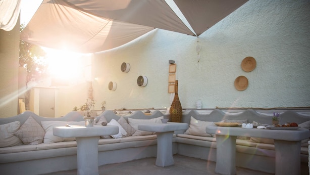 A sunlit outdoor seating area with white sofas and tables, a decorative wall with circular patterns, and a large umbrella providing shade. - Pasithei Studios (Santorini)