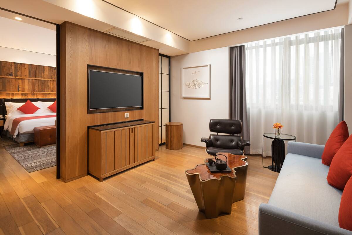 A modern hotel room with a wooden accent wall, a flat-screen TV, a sofa, a coffee table, and a bed with red pillows.