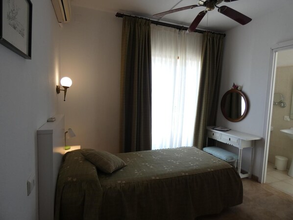 A bedroom with a bed, a ceiling fan, a bedside lamp, a window with curtains, a mirror, and a small vanity with a sink. - Hostal Port Fornells - Adults Only (Mercadal)