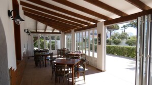 A covered outdoor dining area with wooden beams, tables, and chairs, and large glass doors leading to a garden. - Hostal Port Fornells - Adults Only (Mercadal)