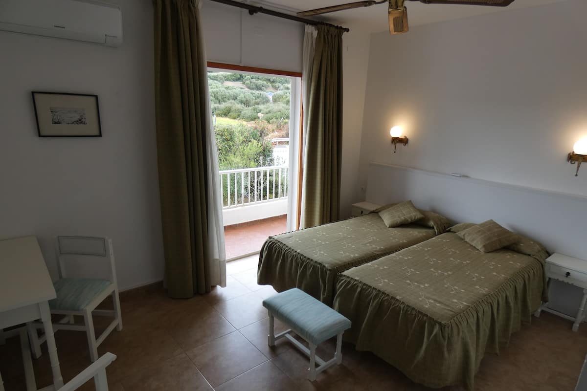 Een slaapkamer met twee bedden, een bankje, een stoel, een nachtkastje, een wandlamp en uitzicht op groen door een raam.