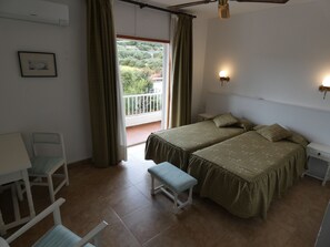 A bedroom with two beds, a bench, a chair, a bedside table, a wall-mounted lamp, and a view of greenery through a window.