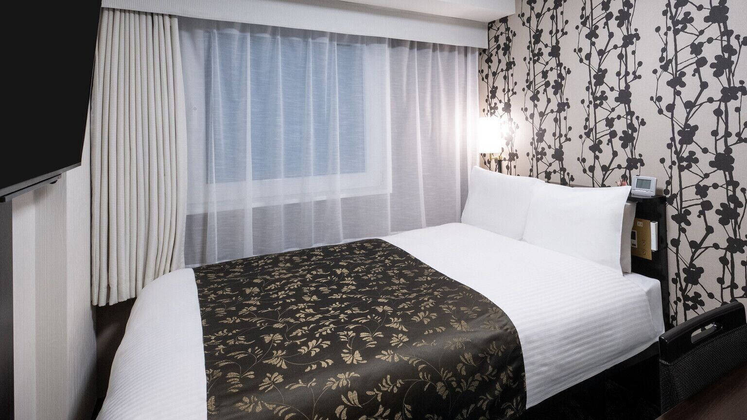 A hotel room with a bed, curtains, a wall with a floral pattern, a bedside table with a lamp, and a TV.