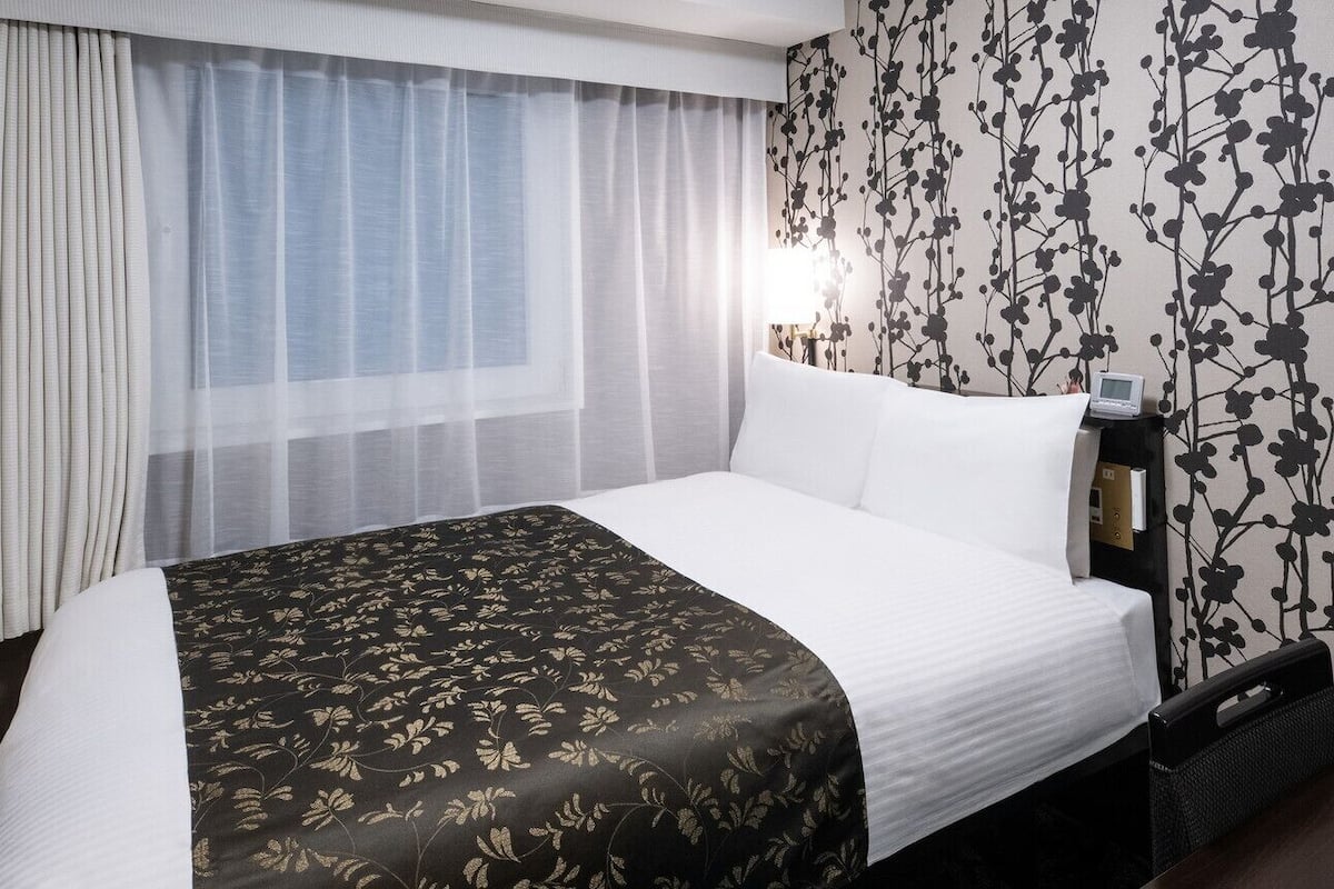 Une chambre d’hôtel avec un lit, des rideaux, un mur orné d’un motif floral, une table de chevet avec une lampe et un téléviseur.