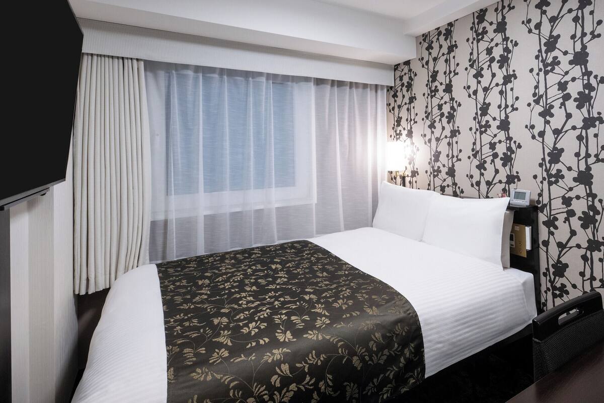 A hotel room with a bed, curtains, a TV, a desk with a lamp, and a floral wallpaper.