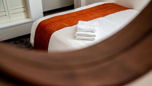 A neatly made bed with a white and orange bedspread, a folded white towel, and a window with white blinds in the background. - Kujira 302 (Osaka)