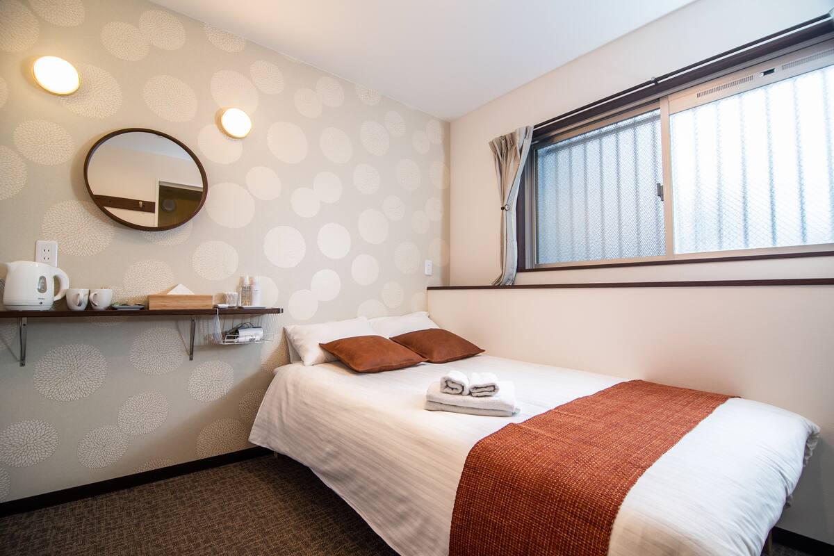 A neatly made bed with a white and brown blanket, a round mirror on the wall, and a window with curtains.
