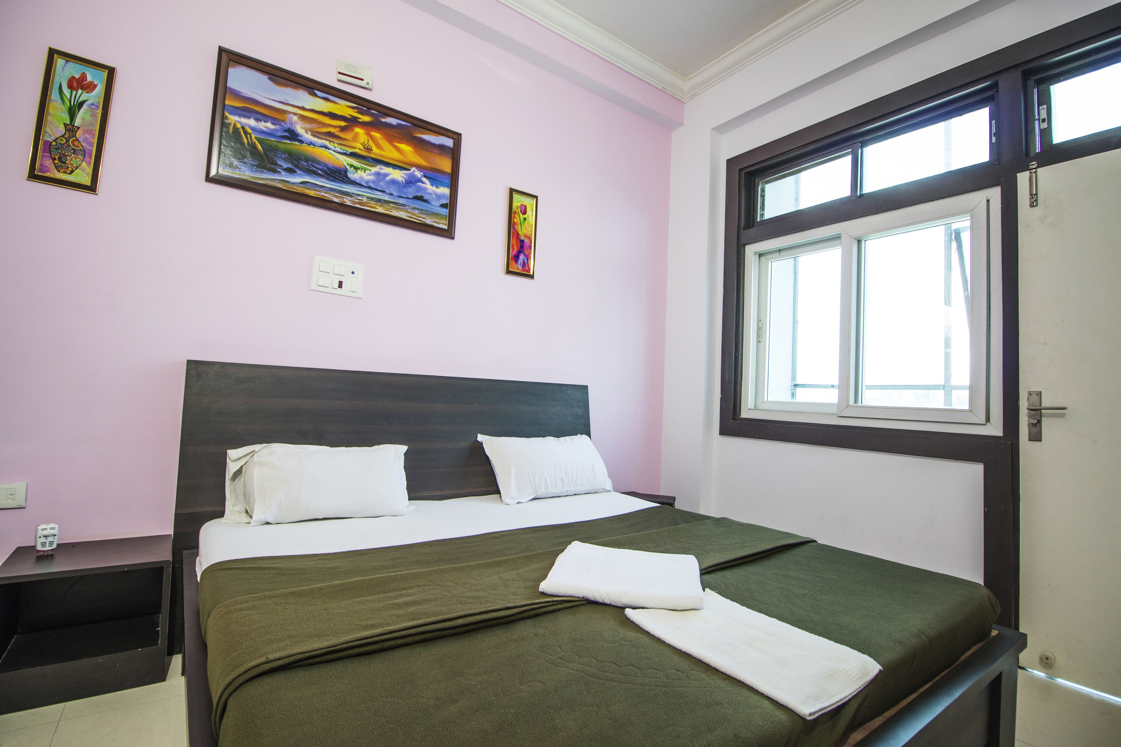 Een net opgemaakt bed met witte kussens en een groene sprei, een nachtkastje met een telefoon, en twee schilderijen aan de muur.