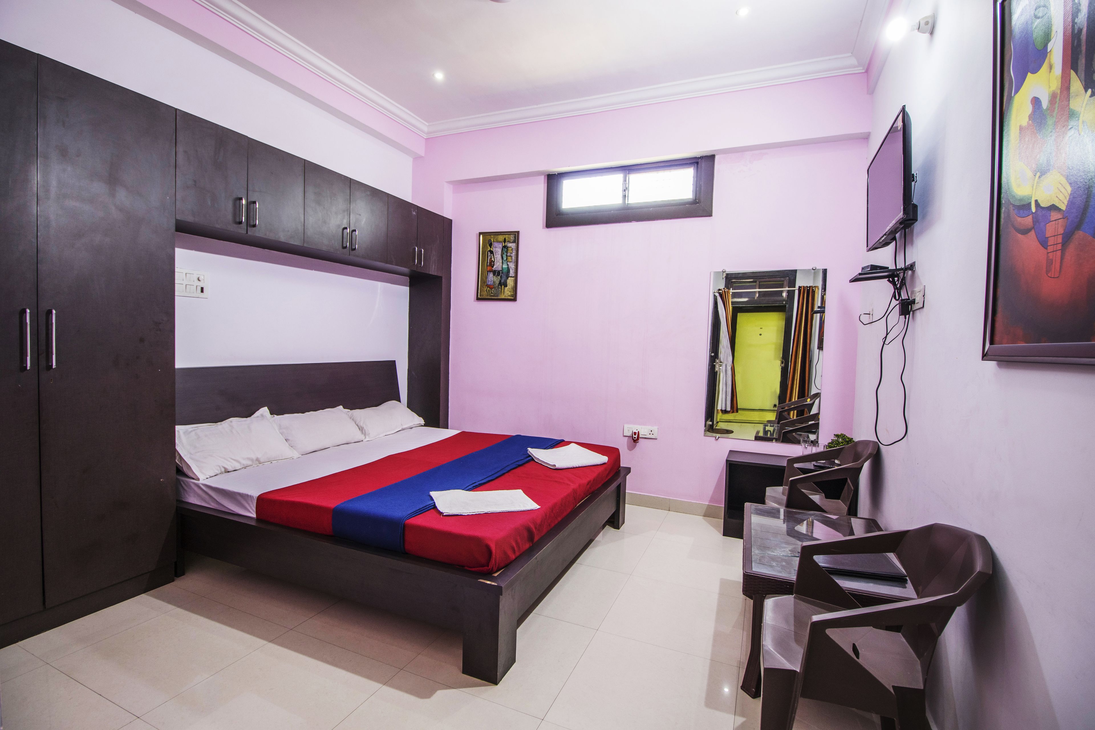 A bedroom with a bed, wardrobe, desk, chair, and a TV on the wall.