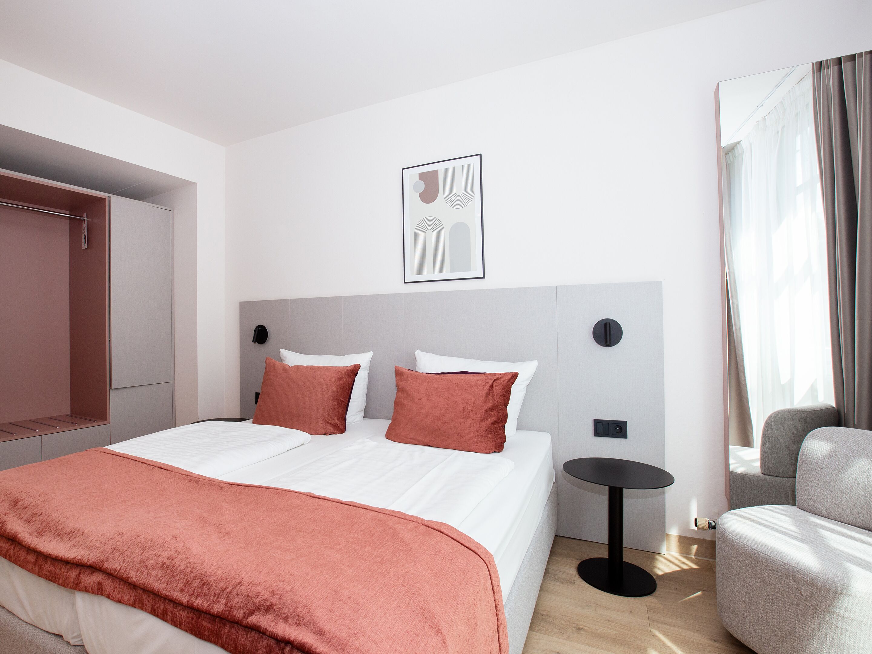 A modern hotel room with a large bed, a bedside table, a chair, and a wardrobe.
