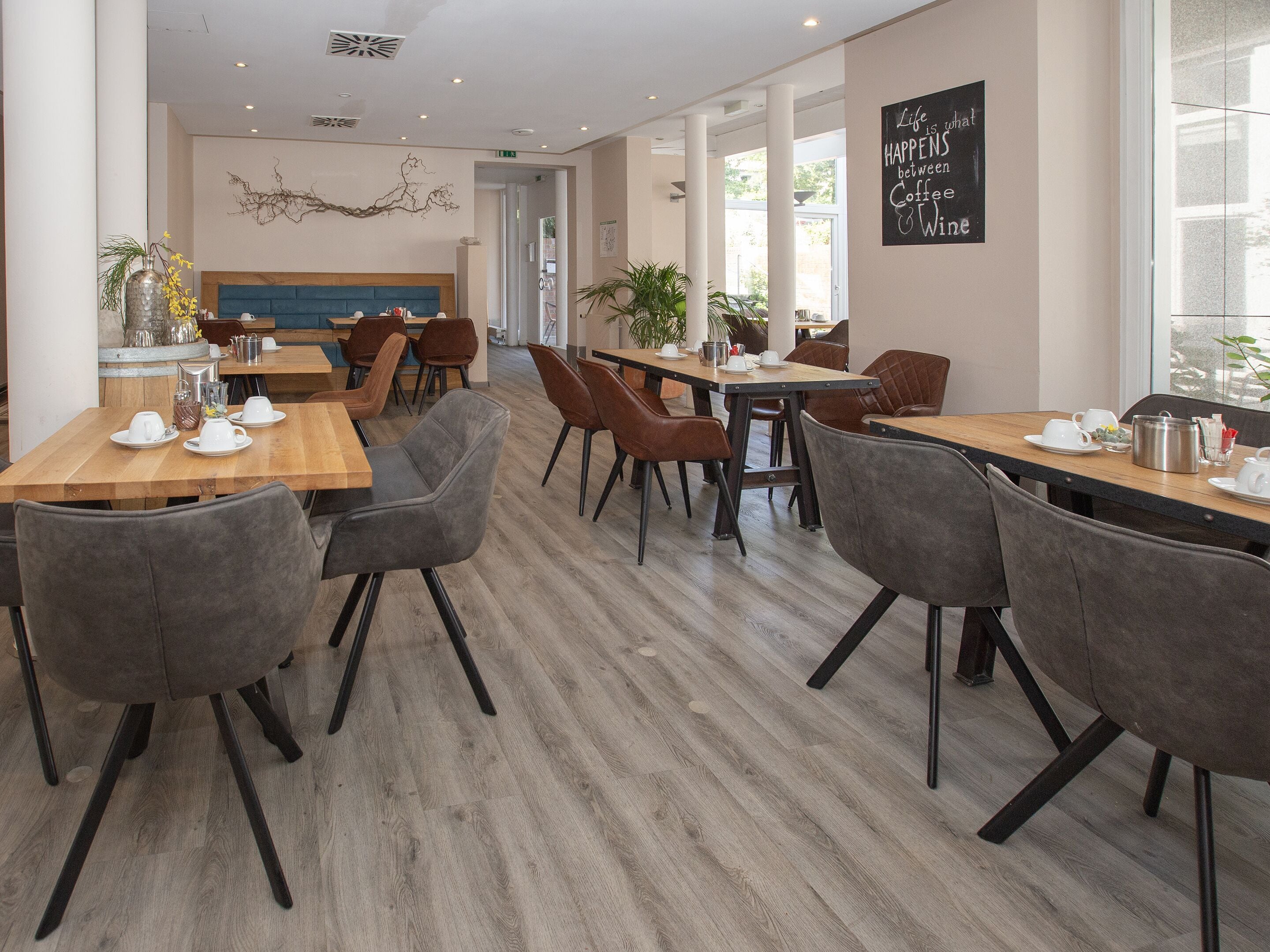 Una cafetería moderna con mesas de madera y sillas grises, que presenta un arte decorativo en la pared y una cita motivacional.