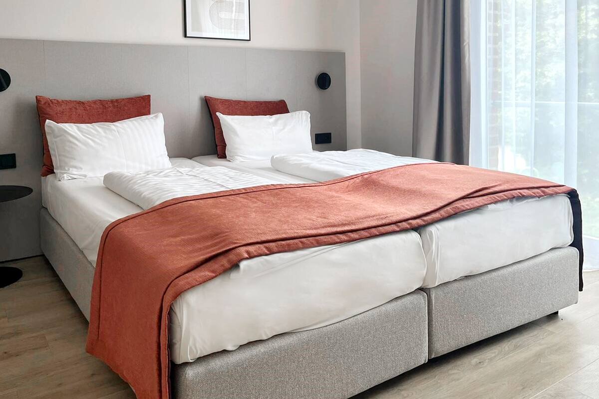 A modern hotel room with a large bed, grey headboard, white bedding, and a view of the outdoors through large windows.