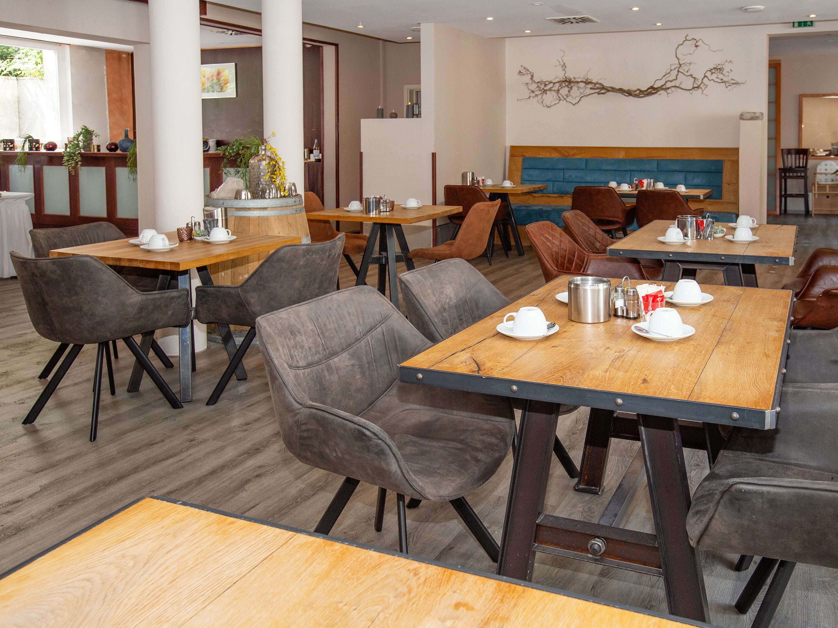 A modern dining area with wooden tables and grey upholstered chairs, featuring a blue cushioned bench in the background.