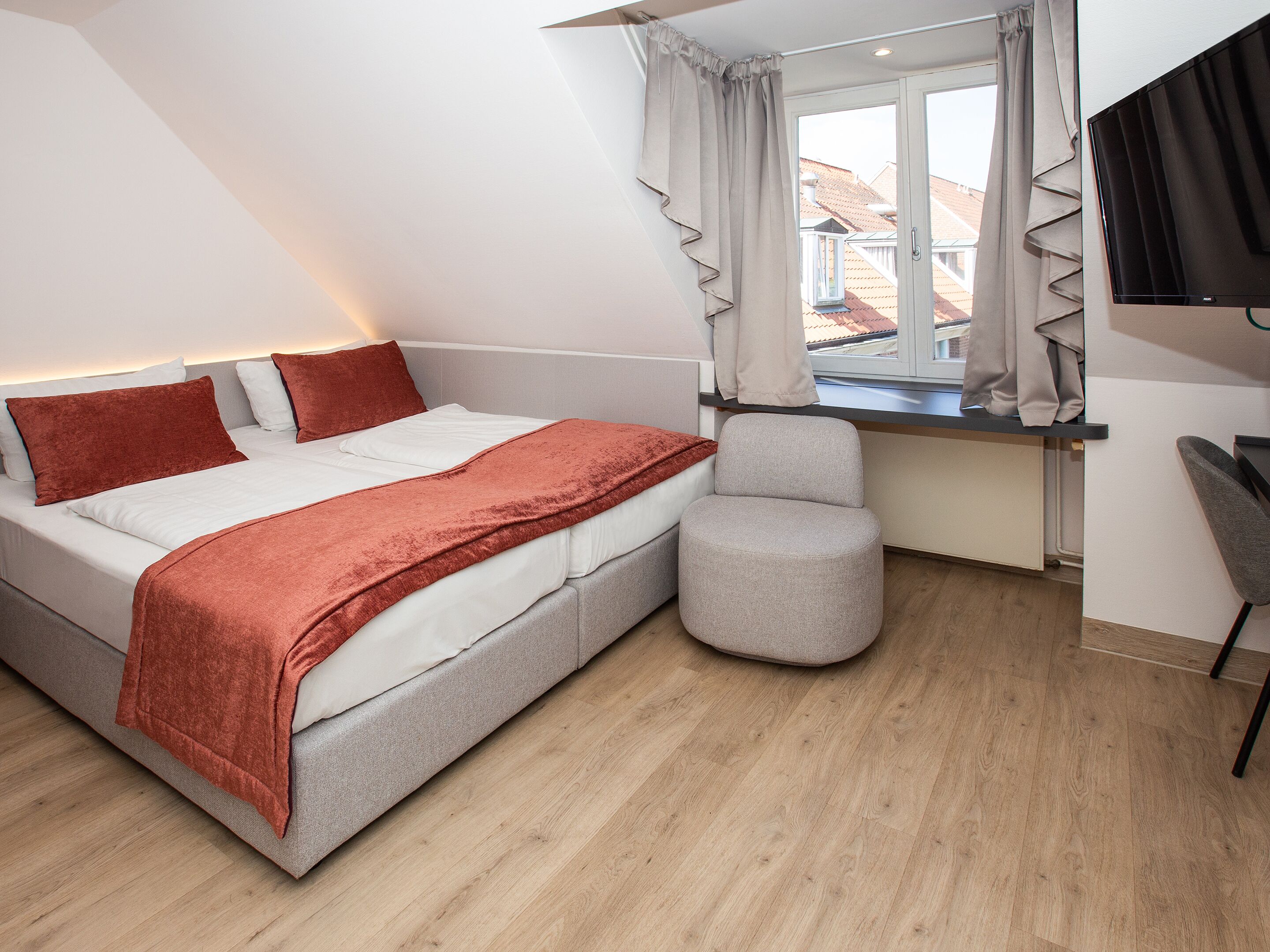 A modern bedroom with a bed, a grey armchair, a window with curtains, and a TV mounted on the wall.