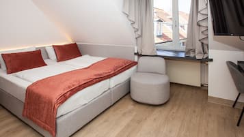 A modern bedroom with a bed, a grey armchair, a window with curtains, and a TV mounted on the wall.