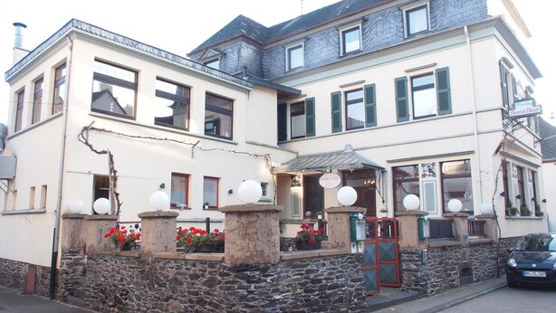 Ein zweistöckiges Gebäude mit einer Stein-Fassade, zahlreichen Fenstern mit Fensterläden und einer kleinen Veranda mit Topfpflanzen und kugelförmigen Dekorationen.