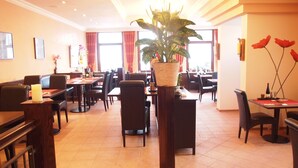Ein Restaurant mit mehreren Tischen und Stühlen, einer großen Kübelpflanze und roten Vorhängen.