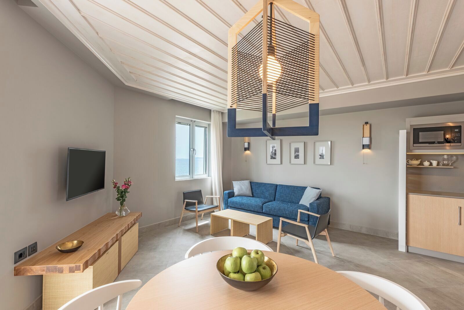Uma sala de estar moderna com um sofá azul, uma mesa de jantar de madeira com uma tigela de maçãs, uma televisão e uma área de cozinha com micro-ondas.