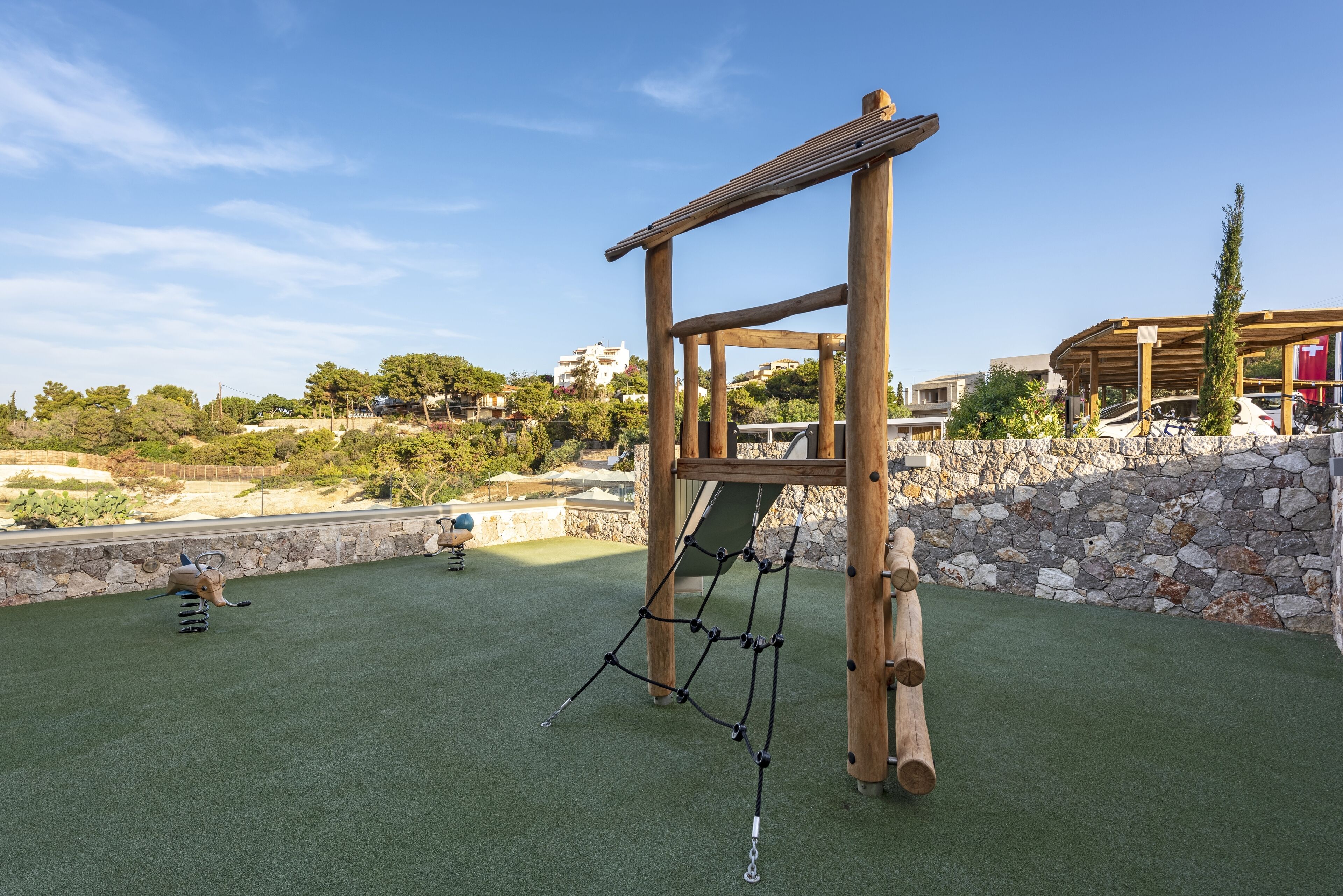 A wooden climbing structure with ropes and a net, set on artificial turf with a stone wall and greenery in the background.