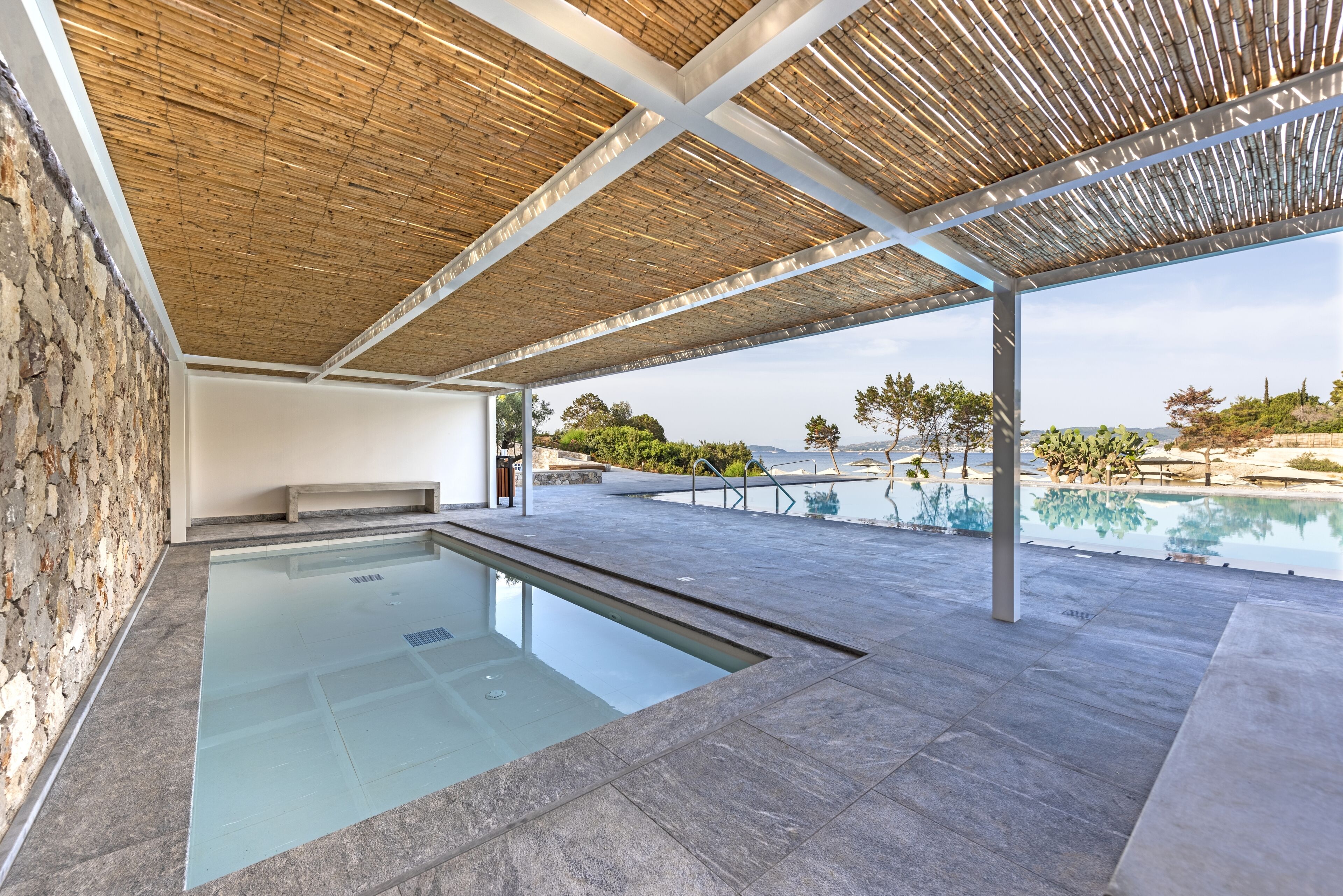 Una piscina moderna al aire libre con un muro de piedra, techo de madera y vistas a la playa y las montañas.