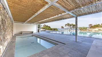 Una piscina moderna al aire libre con un muro de piedra, techo de madera y vistas a la playa y las montañas.