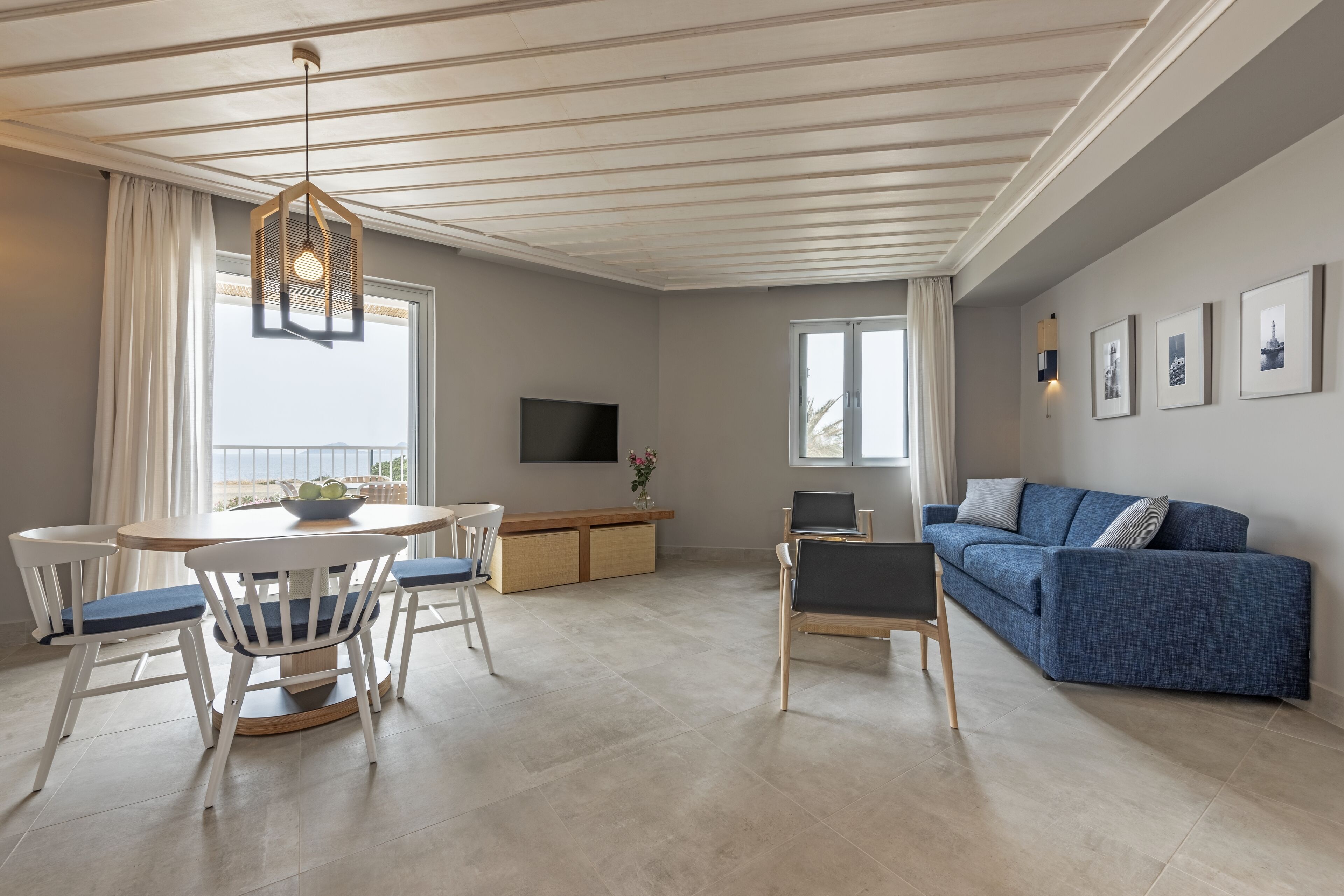 Un soggiorno moderno con un divano blu, un tavolo da pranzo bianco con sedie, una TV a parete e un'ampia finestra con tende.