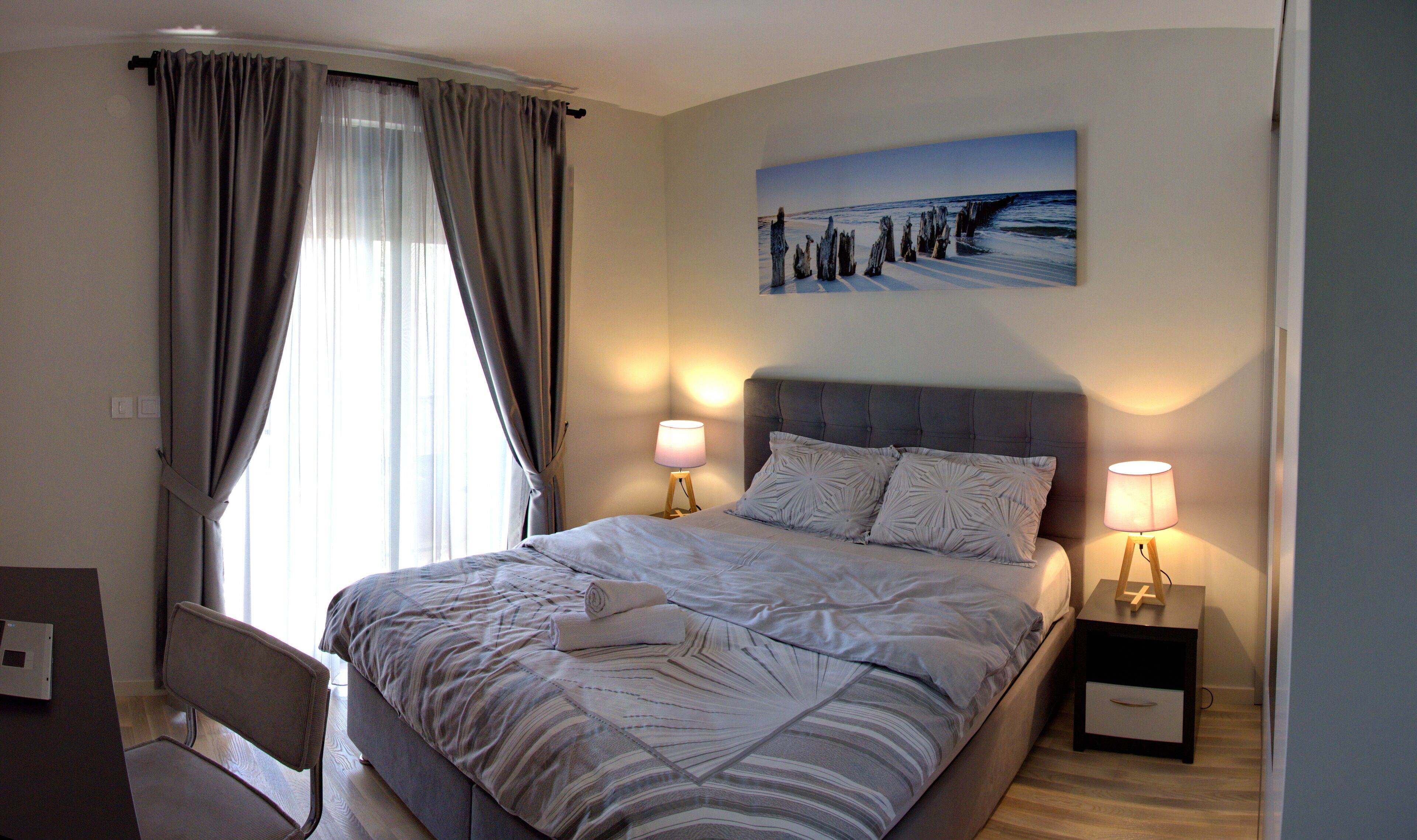 Un lit bien fait, recouvert d’une couvre-lit à rayures, deux lampes de chevet, un tableau représentant un paysage côtier et une chaise dans une pièce dotée d’une grande fenêtre et de rideaux.