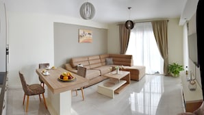 A modern living room with a beige sofa, a wooden dining table with fruit, and a large window with curtains. - Apartmani Ralux Tivat (Tivat)