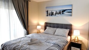 A neatly made bed with a striped bedspread, two bedside lamps, and a painting of a coastal scene on the wall. - Apartmani Ralux Tivat (Tivat)
