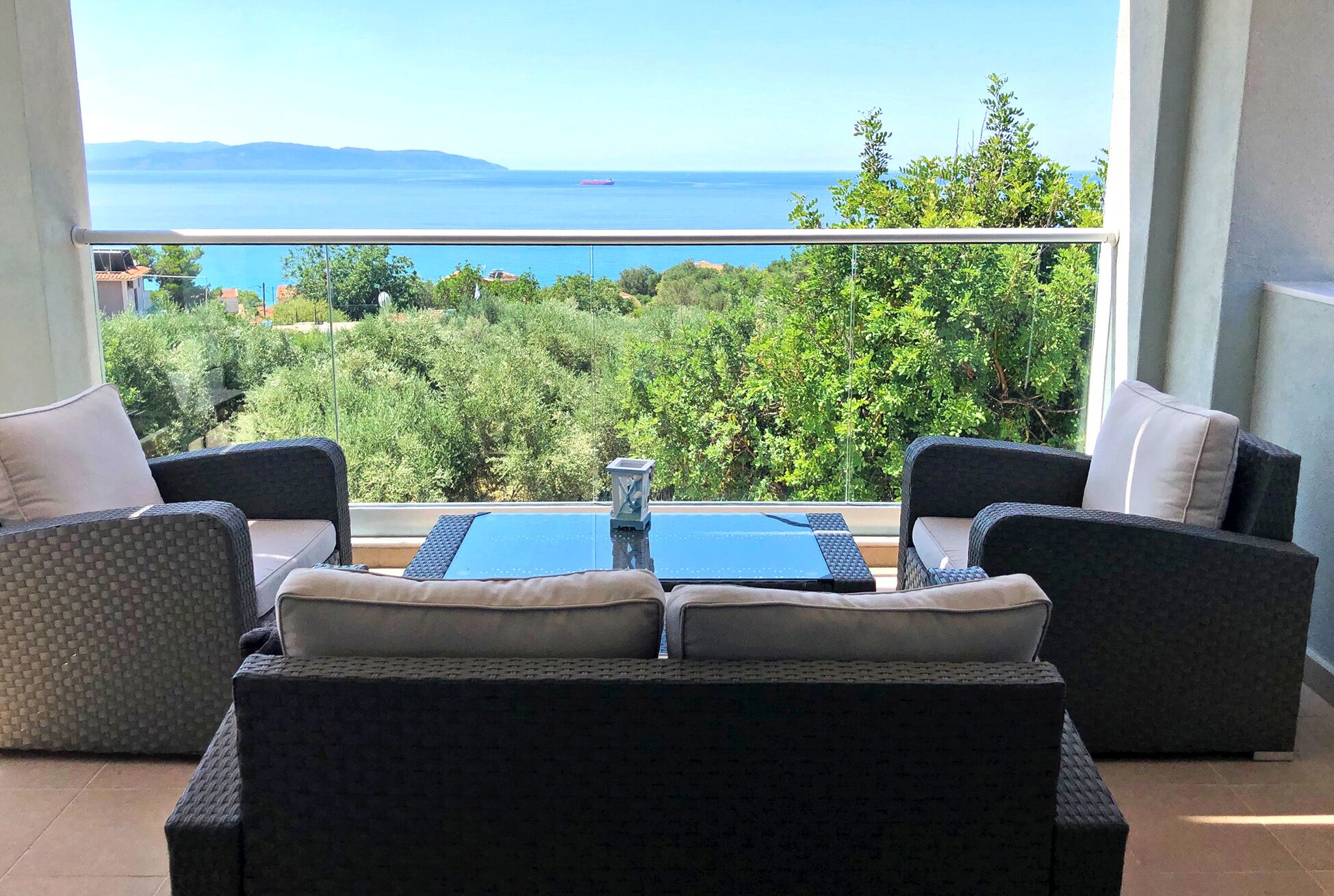 A balcony with wicker furniture, a glass table, and a view of the sea and greenery.