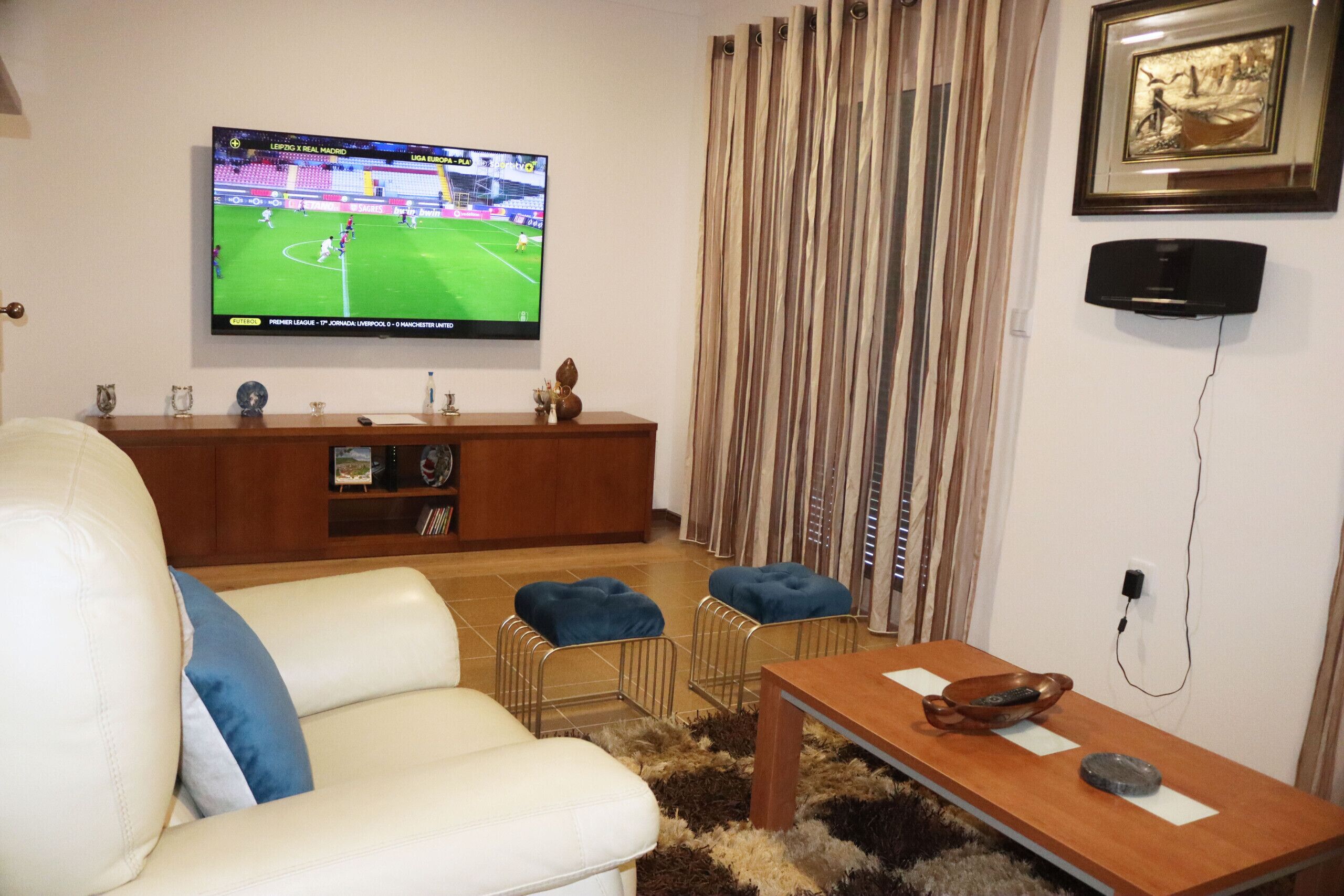 A living room with a flat-screen TV displaying a soccer match, a sofa, a coffee table, and a curtain.