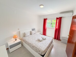 A bedroom with a bed, nightstand, lamp, window with curtains, and an air conditioning unit.