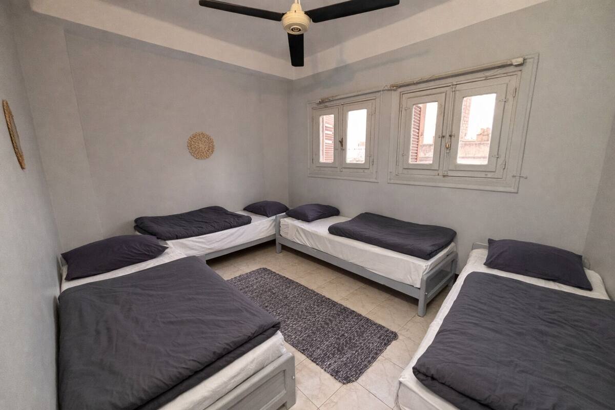 A room with four single beds, a ceiling fan, and a window with shutters.