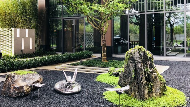 Um edifício moderno com grandes janelas de vidro, uma árvore e um jardim de pedras com uma escultura de pássaro.