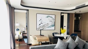 Quarto de hotel moderno com um sofá cinzento, uma mesa de jantar com cadeiras, uma pequena mesa branca e um quadro abstrato grande na parede.